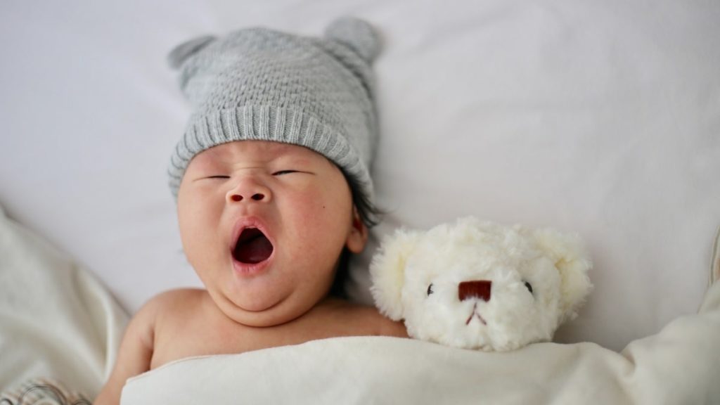 Gapende baby in bed met grijs mutsje op en wit beertje ernaast