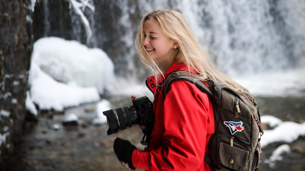 een lachend persoon bij een waterval draagt een camerarugzak en heeft een camera in de hand