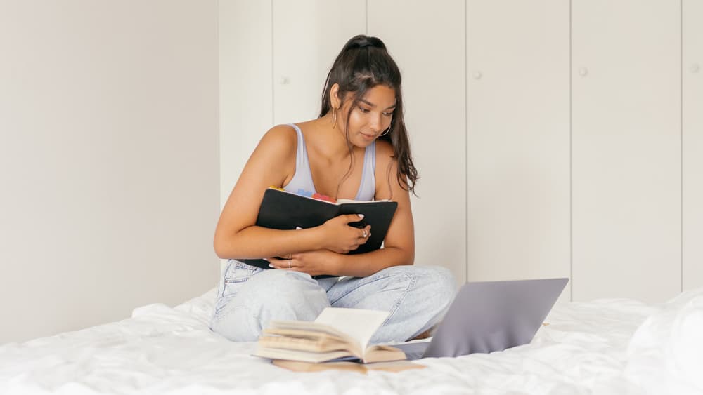 Student zit met laptop en boeken op bed 